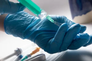 Profissional com luvas azuis manipulando seringa com solucao verde conectada a fixador de cateter para acesso venoso