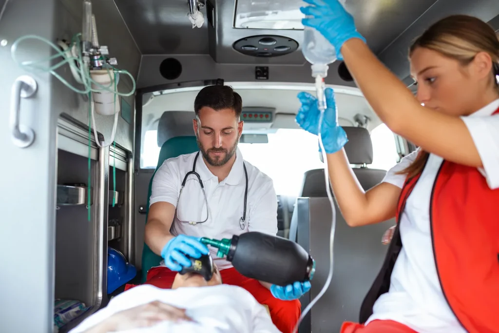 Dois socorristas realizando atendimento pre-hospitalar dentro de ambulancia com oxigenio e soro para paciente em maca
